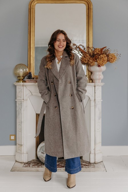 Femme debout qui pose devant un miroir et porte un manteau long gris.